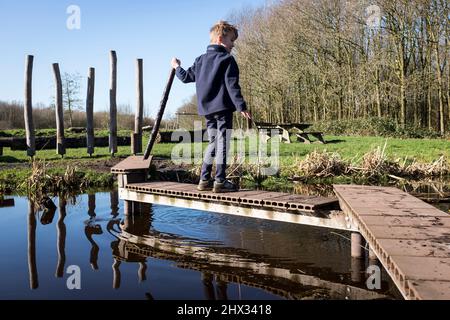 Un jeune garçon joue avec un bâton sur une corniche de bois au-dessus d'un petit canal dans un parc, sous le soleil de la source lumineuse, dans une forêt à Diemen, aux pays-Bas. Banque D'Images