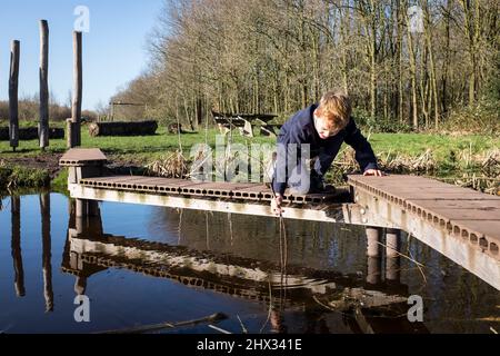 Un jeune garçon joue sur une corniche en bois au-dessus d'un petit canal dans un parc, sous le soleil de la source lumineuse, dans une forêt à Diemen, aux pays-Bas. Banque D'Images