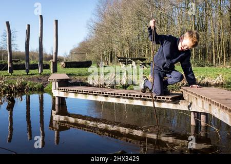 Un jeune garçon joue sur une corniche en bois au-dessus d'un petit canal dans un parc, sous le soleil de la source lumineuse, dans une forêt à Diemen, aux pays-Bas. Banque D'Images