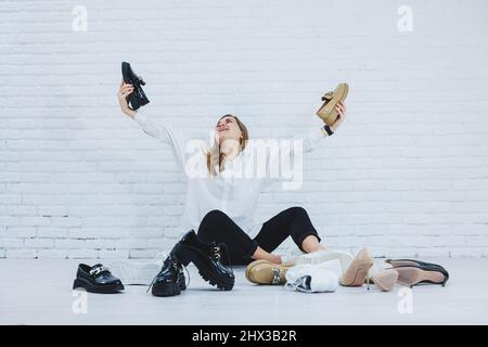 Une jeune femme heureuse avec un sourire dans une tenue tendance avec une chemise blanche et un pantalon noir se trouve sur le sol parmi les chaussures et choisit un Banque D'Images