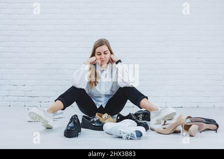 Élégant blond choisissant des chaussures tout en étant assis sur le sol. Vue latérale d'un grand nombre de chaussures pour femmes sur le sol, un choix de chaussures élégantes. Banque D'Images