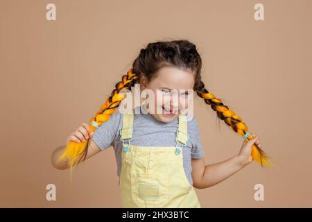 Euphoric petite fille tenant des queues de kanekalon jaunes avec les mains, regardant l'appareil-photo se livrer et sourire avec dent manquante porter jaune Banque D'Images