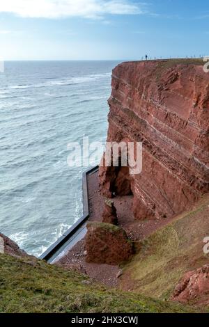 Homme debout sur le bord de hautes falaises rouges spectaculaires de l'île d'Heligoland avec mer rugueuse. Journée d'hiver ensoleillée et venteuse à Helgoland dans la mer du Nord Banque D'Images