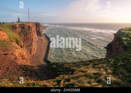 De hautes falaises rouges spectaculaires de l'île d'Heligoland avec une mer rugueuse et un phare loin à l'arrière. Journée d'hiver ensoleillée et venteuse à Helgoland dans la mer du Nord Banque D'Images