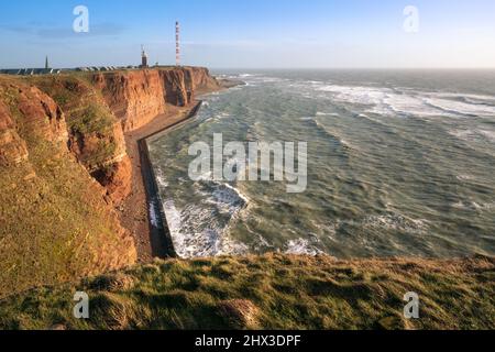 De hautes falaises rouges spectaculaires de l'île d'Heligoland avec une mer rugueuse et un phare loin à l'arrière. Journée d'hiver ensoleillée et venteuse à Helgoland dans la mer du Nord Banque D'Images