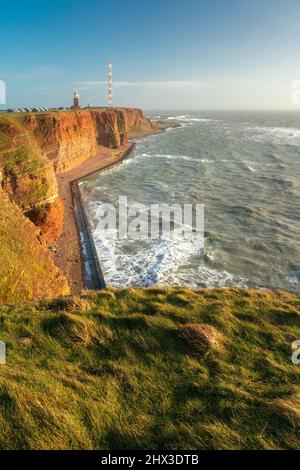 De hautes falaises rouges spectaculaires de l'île d'Heligoland avec une mer rugueuse et un phare loin à l'arrière. Journée d'hiver ensoleillée et venteuse à Helgoland dans la mer du Nord Banque D'Images