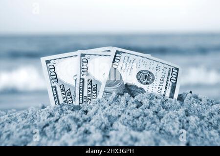 Trois billets de cent dollars à moitié ensevelis dans le sable sur un bord de mer sablonneux Banque D'Images