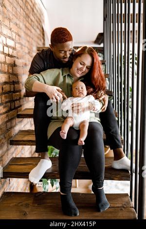 Une famille souriante s'assoit dans les escaliers. Belle mère et père souriant tenant bébé garçon. Joyeux parents regardant doucement leur fils passent les week-ends à h Banque D'Images