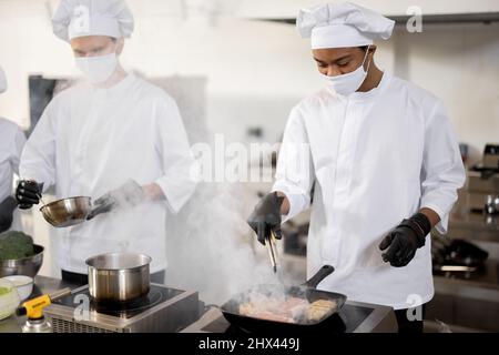 Deux chefs en uniforme et masques de visage cuisinant ensemble dans une cuisine professionnelle. Latin Guy friture de viande et européen homme faisant la pause. Travail acharné au restaurant. Protection en cas de pandémie Banque D'Images