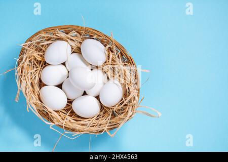 Œufs de pâques blancs dans un panier jaune avec paille sur fond bleu. Vue de dessus du concept de carte postale joyeuses Pâques Banque D'Images