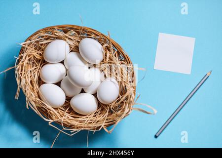 Œufs de pâques blancs dans un panier jaune avec paille sur fond bleu. Vue de dessus du concept de carte postale joyeuses Pâques Banque D'Images