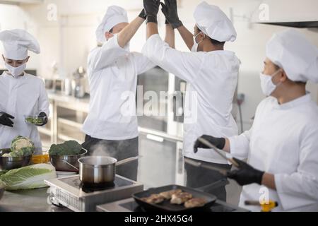 Équipe multiraciale de cuisiniers donnant cinq avec les mains l'une de l'autre, tout en cuisinant dans la cuisine professionnelle. Concept de travail d'équipe et de réussite dans le secteur de la restauration. Cuisiniers portant des masques et des gants de protection Banque D'Images