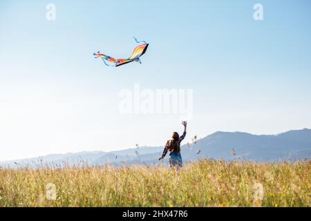 Fille à poil long avec volant un cerf-volant coloré sur la prairie haute herbe dans les champs de montagne. Moments d'enfance heureux ou concept de dépense de temps en extérieur Banque D'Images