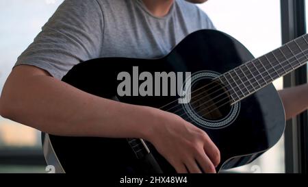 Homme debout sur le balcon contre les fenêtres et jouant de la guitare. Jeune homme passant du temps à la maison pratiquant en jouant de la guitare acoustique, social distanci Banque D'Images