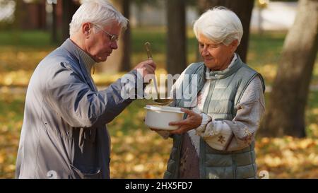 Généreux homme caucasien âgé à poil gris bénévoles dans une organisation caritative et verse de la soupe chaude dans le bol blanc de la femme sans abri âgée. Photo de haute qualité Banque D'Images