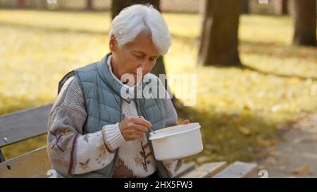 Une femme caucasienne âgée, sans abri, est assise sur un banc à l'extérieur et mange de la soupe chaude qui lui a été donnée par un groupe de bénévoles. Photo de haute qualité Banque D'Images
