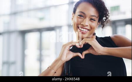 Faire ce que j'aime et aimer ce que je fais. Photo courte d'une jeune femme d'affaires montrant un signe de coeur tout en marchant dans un bureau moderne. Banque D'Images