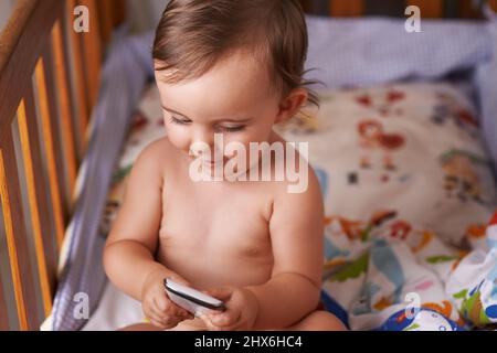 Shes fascinés par le téléphone portable. Une petite fille assise dans son berceau jouant avec son téléphone portable. Banque D'Images