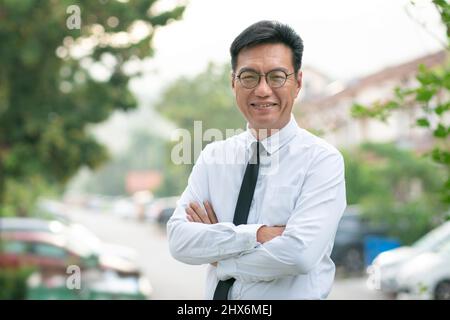 Homme d'affaires asiatique mûr et bien habillé avec les bras croisés, souriant avec assurance. La propriété résidentielle urbaine à l'arrière-plan. Banque D'Images