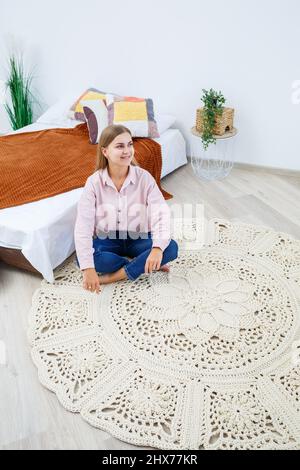 Une femme repose sur un tapis près du lit, un tapis maison en maille de fils épais, fait à la main: Un tapis fait à la main en fils beiges repose sur la fl Banque D'Images