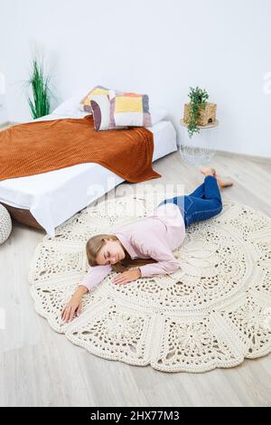 Une femme repose sur un tapis près du lit, un tapis maison en maille de fils épais, fait à la main: Un tapis fait à la main en fils beiges repose sur la fl Banque D'Images