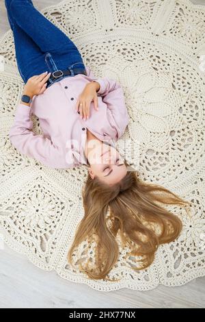 Une femme repose sur un tapis près du lit, un tapis maison en maille de fils épais, fait à la main: Un tapis fait à la main en fils beiges repose sur la fl Banque D'Images