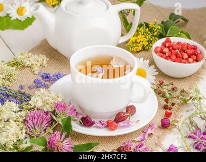 Tisane au rosehip, camomille et trèfle dans une tasse blanche sur une table blanche en bois avec des fleurs. Banque D'Images