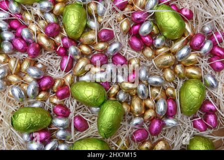 Pile de feuilles pastel colorées de chocolat emballé oeufs de pâques dans le vert, rose, jaune, argent et or, sur nid de crème pané pâle. Banque D'Images