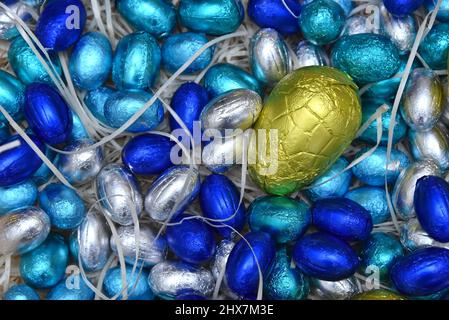 Pile de feuilles pastel colorées, enveloppée de chocolat, œufs de pâques en bleu, argent et turquoise avec un gros œuf jaune au milieu, sur fond pâle. Banque D'Images