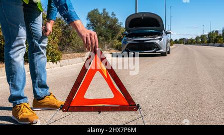 Voiture avec des problèmes et l'homme a mis un triangle rouge pour avertir les autres usagers de la route. Homme conducteur debout près d'une voiture brisée avec capot ouvert, en attente d'aide. Banque D'Images
