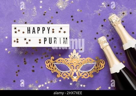 Happy Purim écrit en visionneuse, champagne et masque de carnaval doré sur fond lilas. Concept de fête du carnaval de Purim. Vue de dessus Banque D'Images