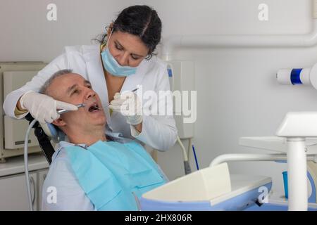 L'homme d'âge moyen est assis sur une chaise de dentiste avec la bouche ouverte tandis que le dentiste travaille sur sa dent malade. Banque D'Images