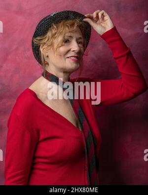 Une femme blanche d'âge moyen est vêtue de rouge et porte une cravate masculine rouge et grise et un chapeau Banque D'Images