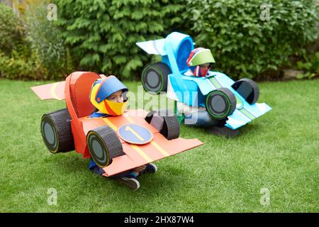 Enfants portant des modèles/costumes de voitures de course en carton, jouant dans le jardin Banque D'Images