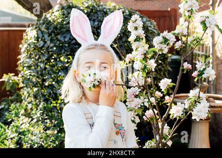 Adorable petite fille blonde dans les oreilles de lapin costume renifler la branche d'arbre en fleur à l'extérieur le jour du printemps. Enfant s'amusant sur la chasse aux œufs de Pâques dans le guar Banque D'Images