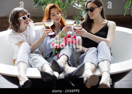 Trois jeunes amies adultes assises avec des boissons alcoolisées dans la baignoire, faisant une fête de poules ou célébrant les vacances des femmes à la maison Banque D'Images
