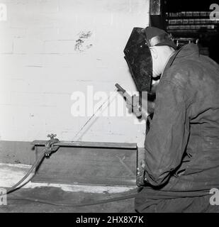 1950s, historique, un travailleur portant un masque facial ou un écran de protection pour usage intensif et des gants utilisant un poste à souder à l'arc électrique sur une plaque métallique sur une petite poutre en acier, Abbey Works, Port Talbot, Neath, pays de Galles, ROYAUME-UNI. Banque D'Images