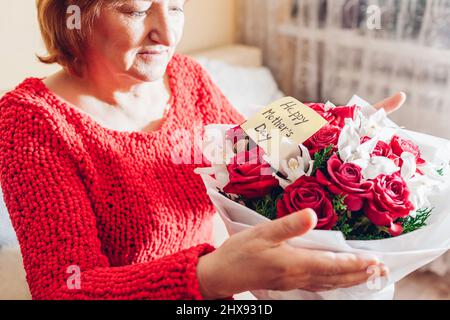 Bonne surprise de la fête des mères. La femme âgée tient un bouquet de roses rouges et des fleurs d'orchidées à la maison avec une carte de vœux. Cadeau pour les vacances de printemps Banque D'Images