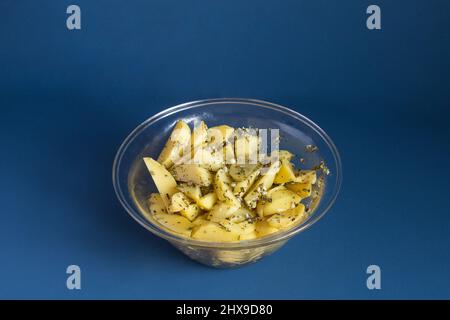 Bol en verre transparent avec pommes de terre crues, sauce à l'ail et romarin prêt à cuire. Une alimentation saine sur fond bleu. Banque D'Images