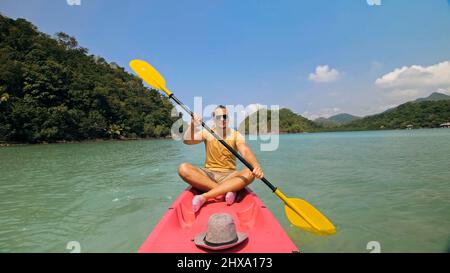 Jeune homme avec des lunettes de soleil et un chapeau rows en plastique rose canoë le long de la mer contre des îles vertes vallonnées avec des jungles sauvages. Voyager dans les pays tropicaux. St Banque D'Images