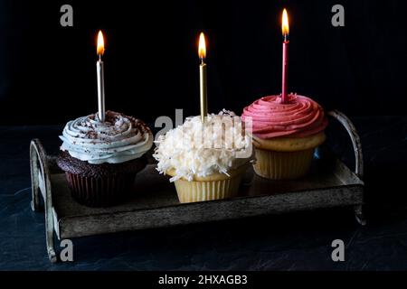 Trois petits gâteaux aux bougies allumées sur un plateau en métal, sur fond sombre. Banque D'Images