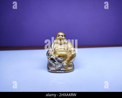 Une figurine dorée de Bouddha riant sur une surface blanche avec un fond violet symbolise la joie, la chance et la prospérité avec sa pose joyeuse et radieuse. Banque D'Images