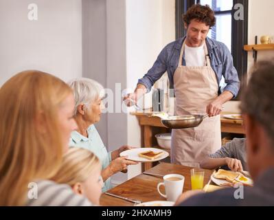 Un seul pour moi, s'il vous plaît. Un petit plan d'un homme servant un petit déjeuner délicieusement cuisiné à sa famille. Banque D'Images