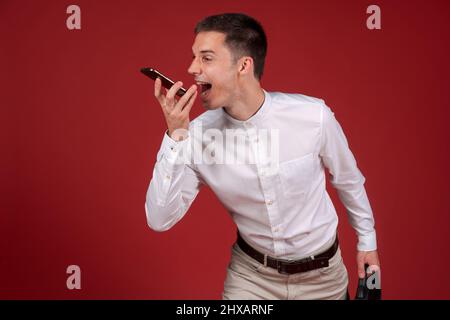 Un homme en colère dans une chemise blanche et un pantalon léger avec un sac à documents dans sa main hurle sur un téléphone portable sur le haut-parleur.Un homme stressé et déprimé sur fond rouge.Homme émotionnel Banque D'Images