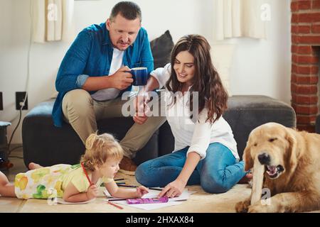 Faire participer tout le monde. Photo courte d'une jeune mère et d'un père aidant leur adorable petite fille à colorer et à dessiner dans le salon à Banque D'Images