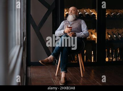 bel homme avec un verre de vin assis dans une chaise près d'un rack de vin de collection. Banque D'Images