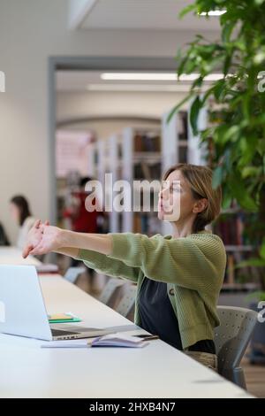 Femme obtenant le deuxième degré de prendre la pause de l'apprentissage tout en étudiant en ligne sur ordinateur portable dans la bibliothèque Banque D'Images