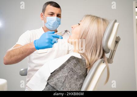 femme en clinique dentaire. Un dentiste en gants bleus en latex enlève sa dent avec un rétracteur buccal et des pinces. Banque D'Images