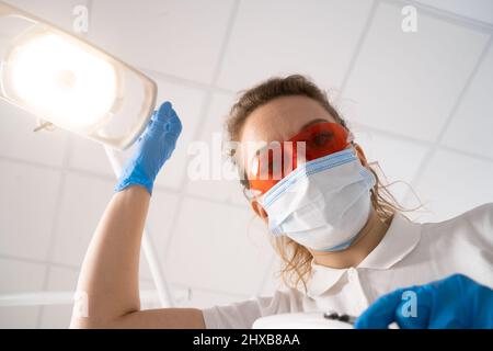 Gros plan d'une jeune femme dentiste en lunettes de protection rouges aux ultraviolets, porte un masque médical, le dentiste traite ses dents. POV du patient. Medic Banque D'Images