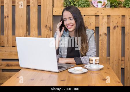 Femme positive spécialiste à distance des vêtements formels assis à table avec du café et du macaron et ayant la conversation sur le téléphone portable tout en travaillant à distance Banque D'Images